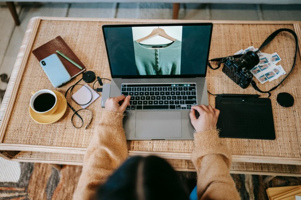 High angle of crop unrecognizable female designer showing photography of apparel on netbook screen at desk with coffee in house